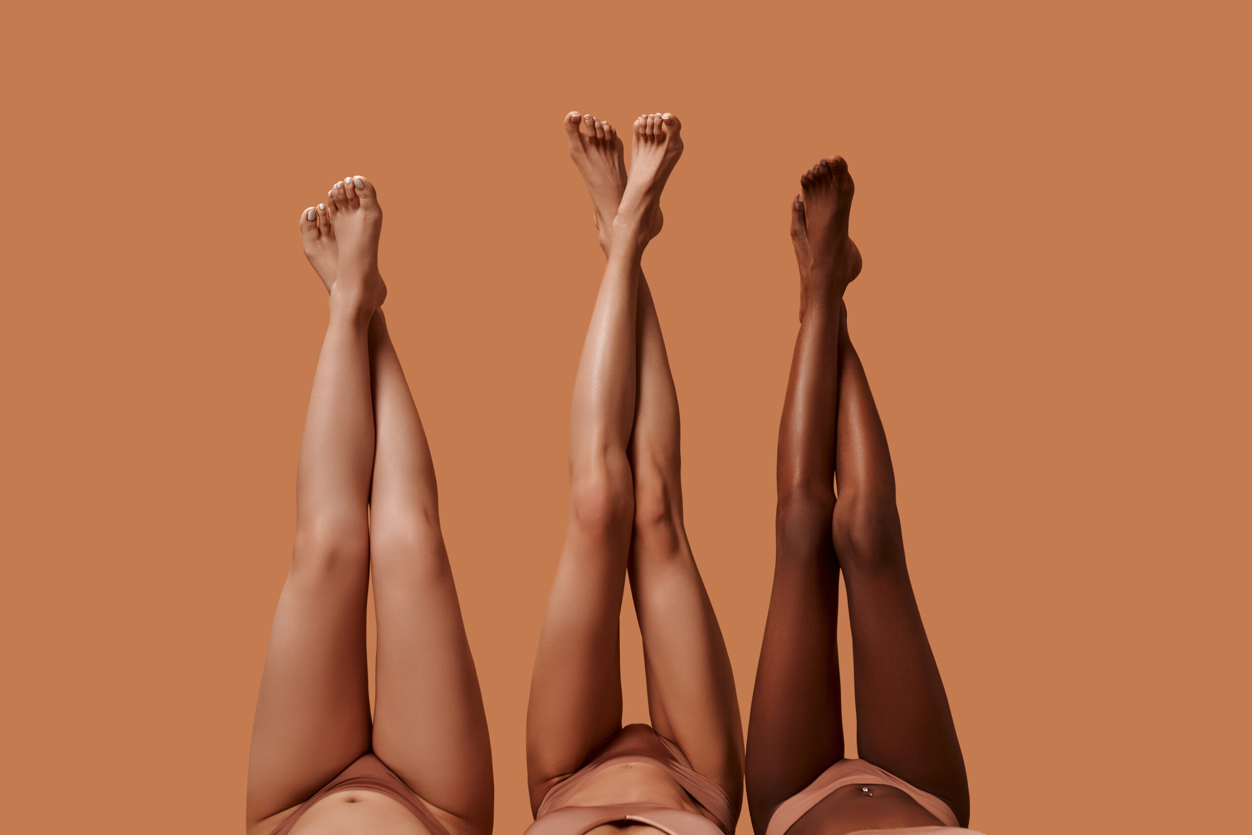 Portrait of beautiful young women of different ethnicities with perfect firm and slim bodies with hairless soft and silky legs crossed isolated on a beige background.